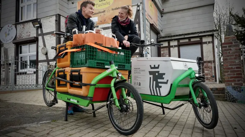 Zwei Ehrenamtliche lachen auf zwei grünen Lastenrädern vor dem Café Tacheles. Die Räder sind mit Warmhalteboxen und Brottüten beladen.